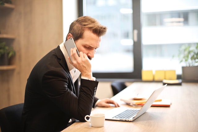 A property manager in a black suit who is receiving an emergency maintenance call from a tenant sits at their desk and arranges repair services on their laptop